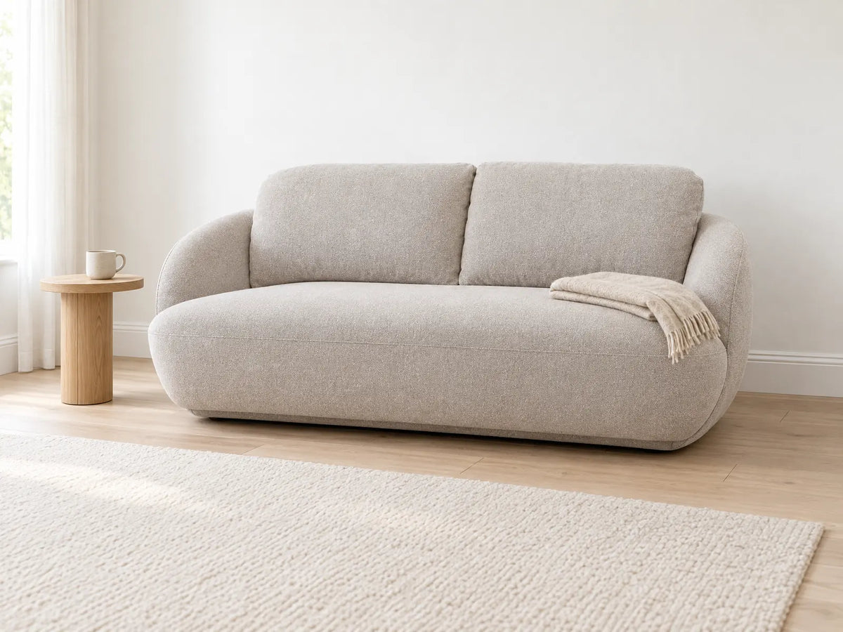 Le canapé banquette bas en coton grège dans un salon lumineux avec un tapis crème, un plaid sur un côté et une table d'appoint ronde en chêne